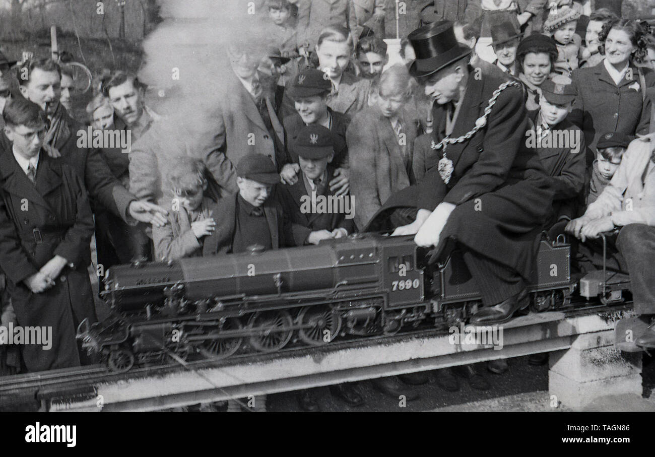 Années 1950, historiques, une foule de personnes regardant un homme âgé d'un dignitaire, un lolca maire portant un haut de forme, et un matin, la chaîne d'avoir un tour sur un train à vapeur miniature, sur un modèle de fer, England, UK. Cet étroit chemin de fer miniature jauge de est relevée au-dessus du sol et le conducteur et les passagers assis à califourchon sur la piste. En cette ère de fer miniature ou modèle avec des locomotives à vapeur ont été une attraction populaire dans les parcs et les villes de bord de mer. Banque D'Images