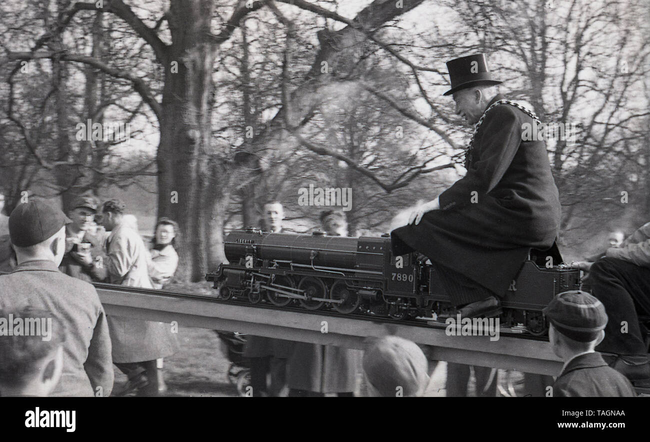 Années 1950, historique, d'un dignitaire local, le maire, portant un haut de forme, et un matin, la chaîne d'équitation une locomotive à vapeur miniature sur un modèle ou un chemin de fer miniature, England, UK. Les étroits chemins de jauge de est relevée au-dessus du sol et le conducteur et les passagers assis à califourchon sur la piste. En cette ère, de modèle ou de chemins de fer miniature avec des locomotives à vapeur ont été une attraction populaire dans les parcs et les villes de bord de mer. Banque D'Images