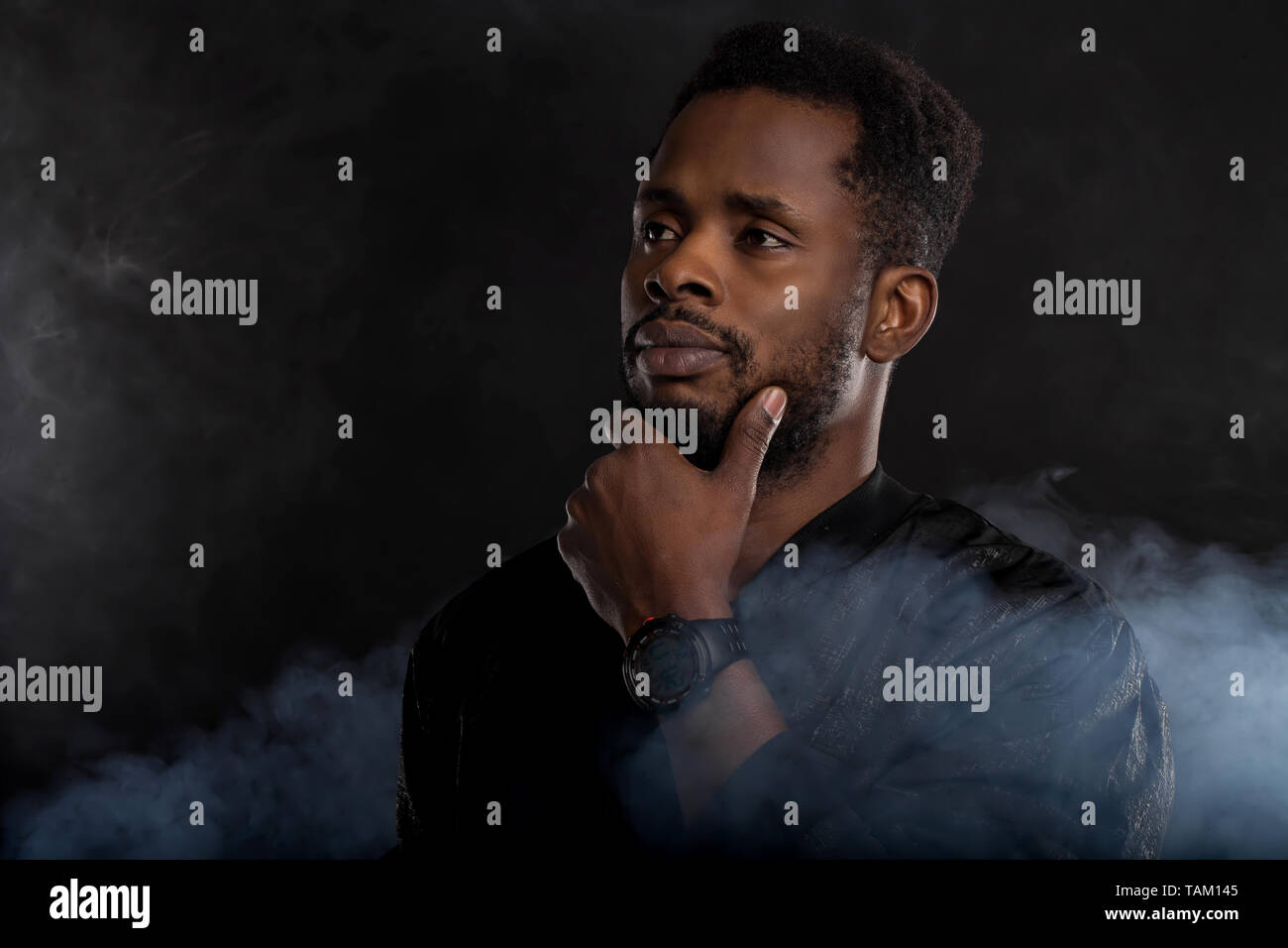 Portrait de teint foncé homme sérieux holding chin, vêtus de noir, veste coupe-vent wristwatces, debout sur un fond sombre avec white smok Banque D'Images