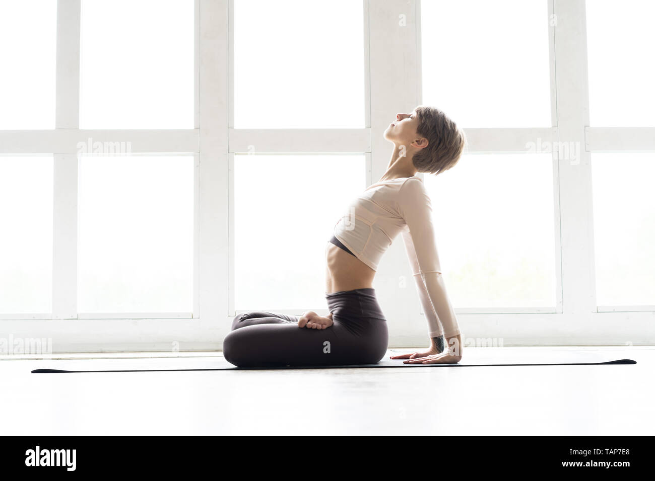 Young attractive smiling woman practicing yoga, assis en demi-lotus, exercice Ardha Padmasana posent, de travail, le port des vêtements de sport Banque D'Images