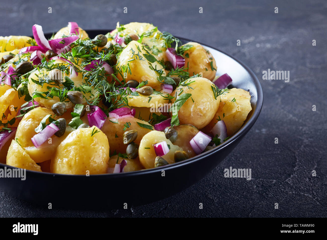 Close-up of Savoury allemand nouveau salade de pommes de terre avec l'oignon rouge, les câpres, les verts et la moutarde de vinaigre dans un bol noir sur une table, la vue à partir de Banque D'Images