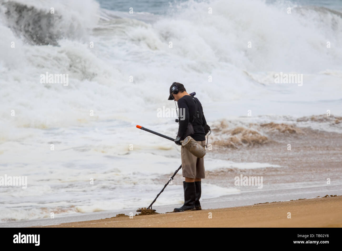 Homme portant des bottes Wellington avec détecteur de métal à la recherche de la côte d'une plage avec des vagues dans l'arrière-plan Banque D'Images