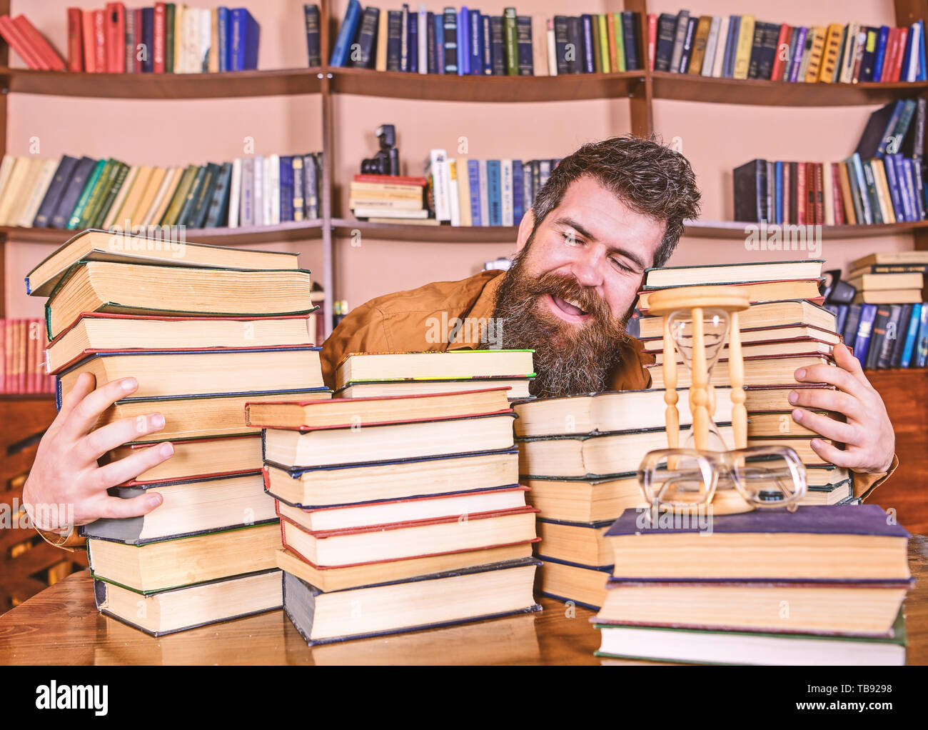 Concept bibliothécaire. Sur l'homme heureux face entre les piles de livres, tout en étudiant en bibliothèque, des étagères sur l'arrière-plan. Enseignant ou étudiant avec barbe est assis à table avec des livres, des flous artistiques. Banque D'Images