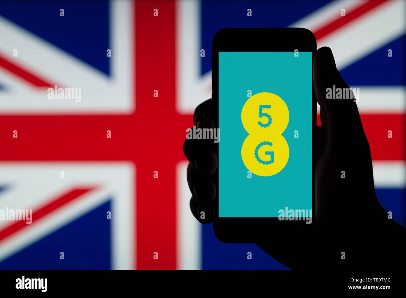 La silhouette d'une main d'un homme est titulaire d'un smartphone affichant le logo de l'ERE 5G, devant un drapeau britannique (usage éditorial uniquement). Banque D'Images
