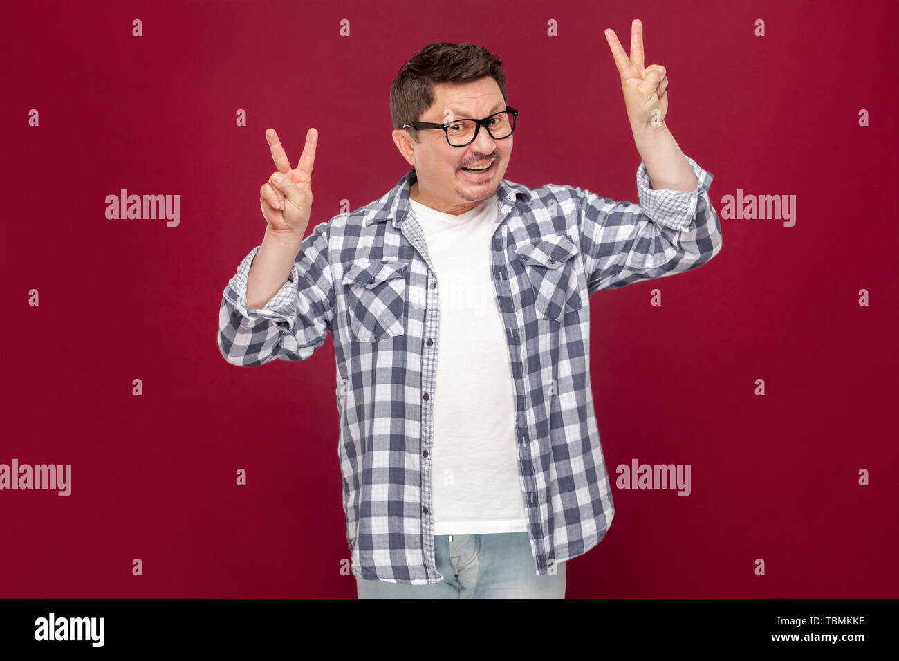 Portrait de funny happy middle aged man in casual chemise à carreaux, les lunettes debout avec la paix ou la victoire et à la caméra à pleines dents avec sm Banque D'Images