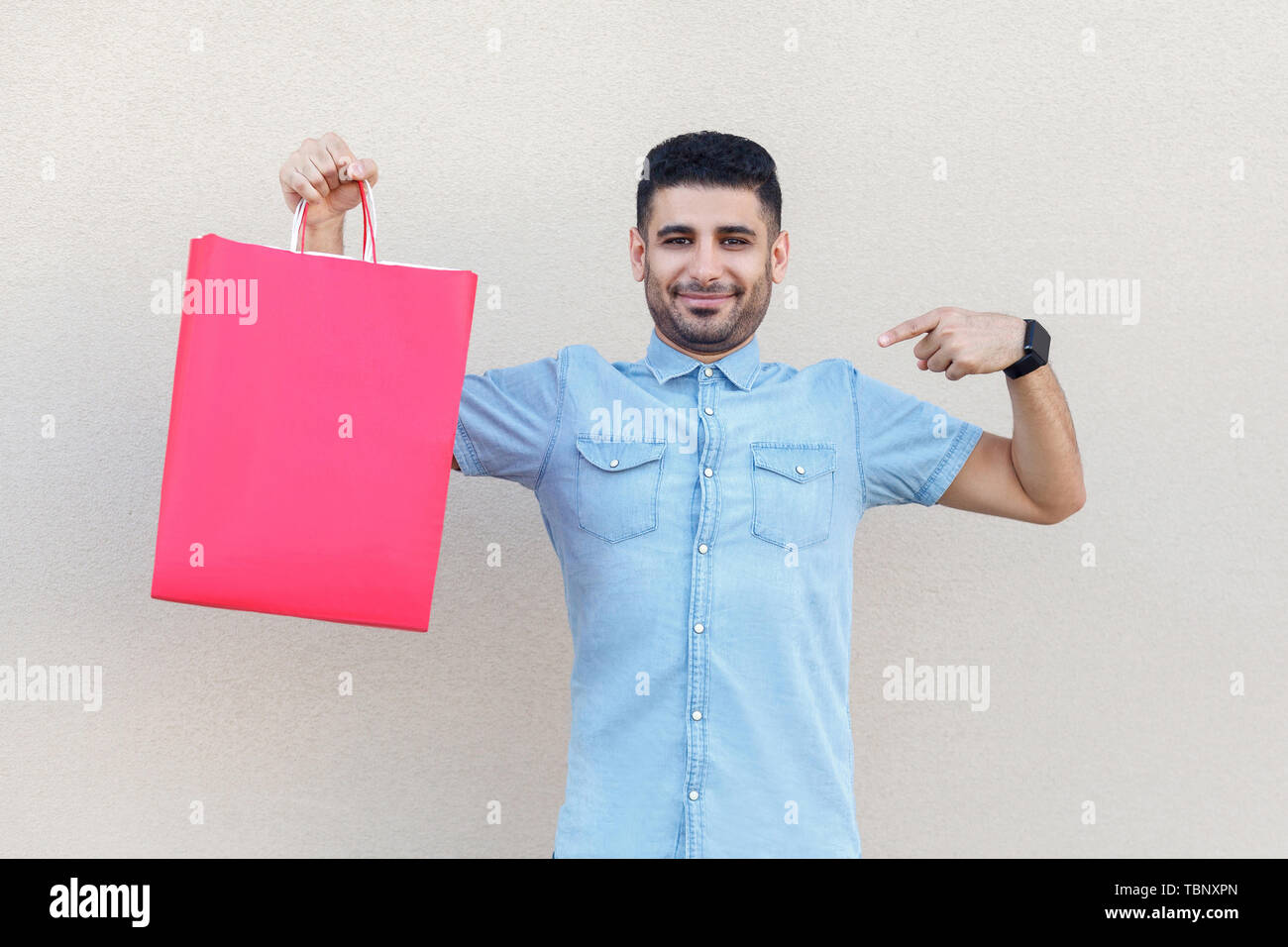 Portrait de beau jeune homme barbu en chemise bleue, tenant debout et pointant au red panier et looking at camera avec sourire. Piscine studi Banque D'Images