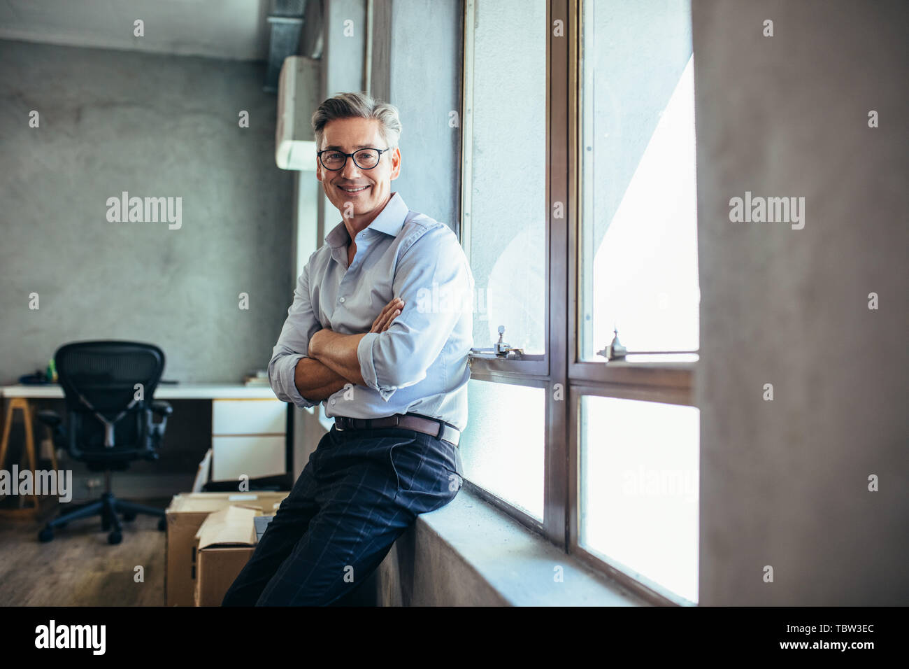 Smiling businessman looking at camera comme il se distingue par un rebord de fenêtre avec les bras croisés. Mid adult businessman à son bureau. Banque D'Images Smiling businessman looking at camera comme il se distingue par un rebord de fenêtre avec les bras croisés. Mid adult businessman à son bureau. Banque D'Images