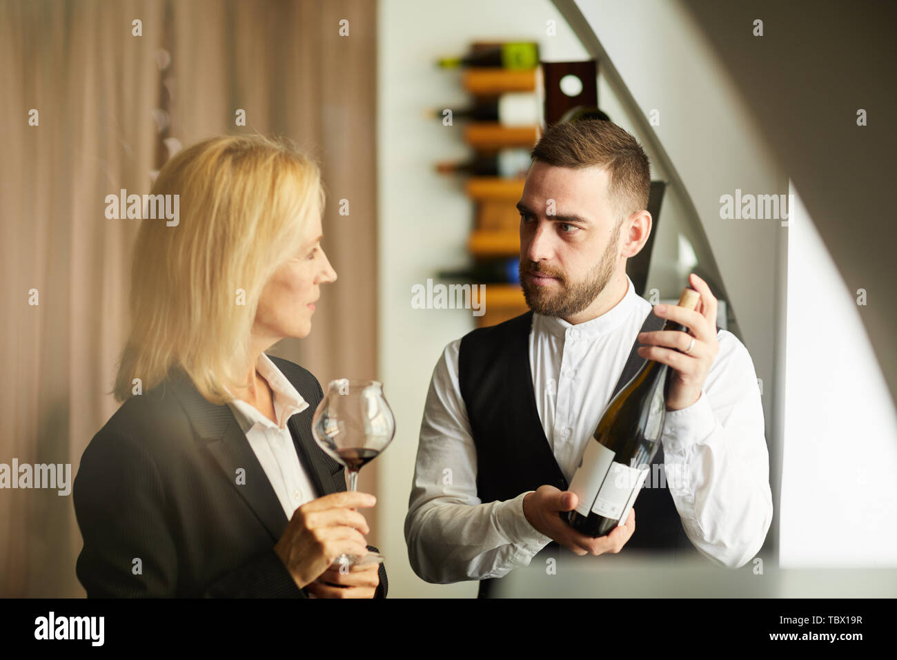 Portrait de sommelier aux teintes chaleureuses montrant la bouteille de vin à la femme au restaurant, copy space Banque D'Images
