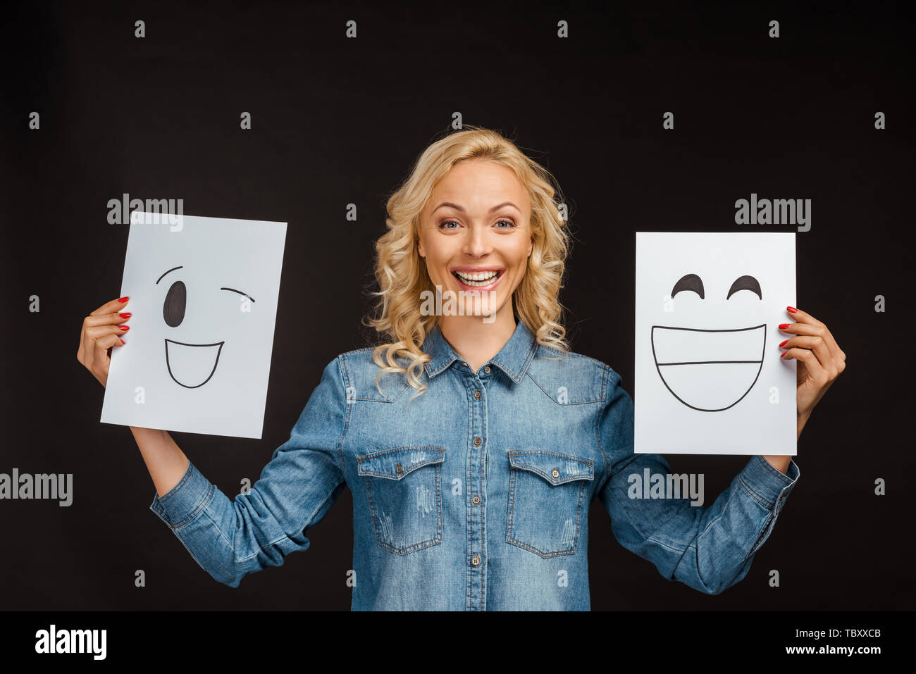 Cheerful blond woman holding les papiers avec des visages heureux isolé sur black Banque D'Images