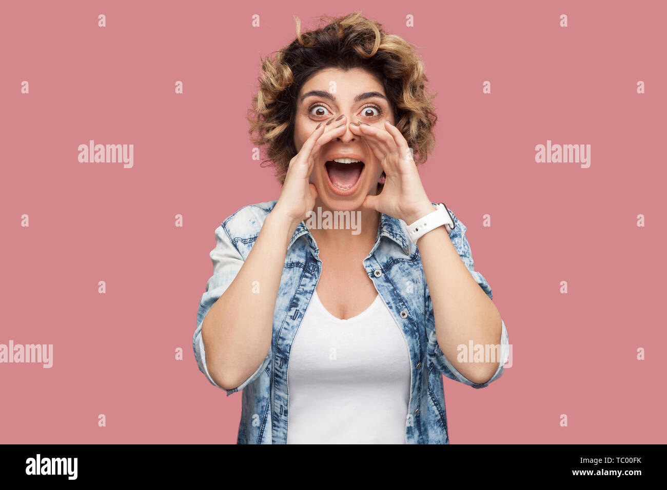 Portrait of young woman with curly hairstyle in casual shirt bleu, debout, criant quelque chose en commun et à la caméra avec étonnement à fac Banque D'Images