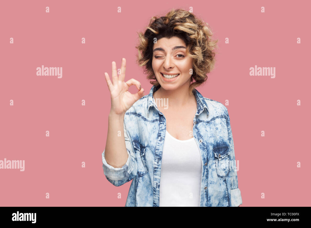 Portrait de jeune femme drôle avec curly hairstyle in casual chemise bleue avec permanent signe ok par contre, geste, sourire à pleines dents et clignant à camer à Banque D'Images