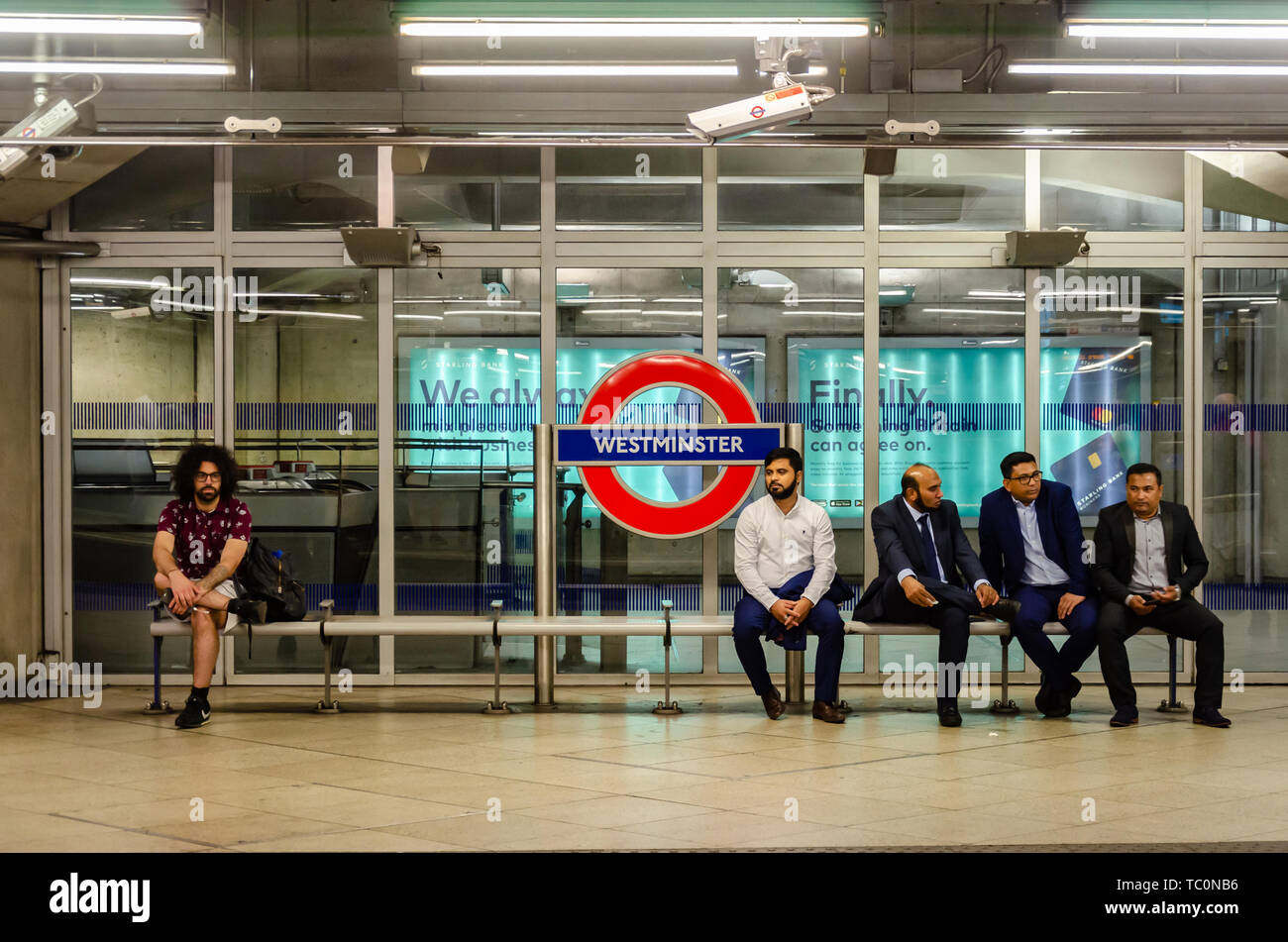 Un homme est assis seul sur un banc sur la plate-forme à la station de métro Westminster comme un groupe d'hommes asiatiques assis ensemble. Banque D'Images