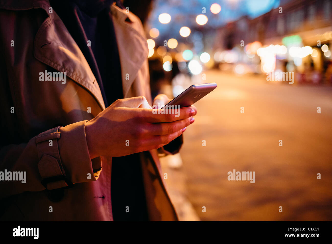 Jeune femme élégante à l'aide de mobile phone debout sur la rue de la ville de nuit éclairée par les lumières de fête chaleureuse, close-up. Banque D'Images