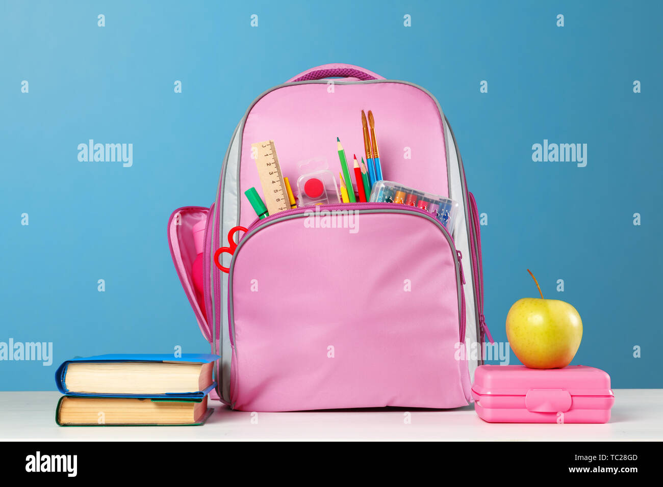 Ensemble des élèves. Sac à dos rose avec modèle, une pile de livres, une  boîte à lunch, une pomme sur la table sur un fond bleu. Retour à l'école  Photo Stock -