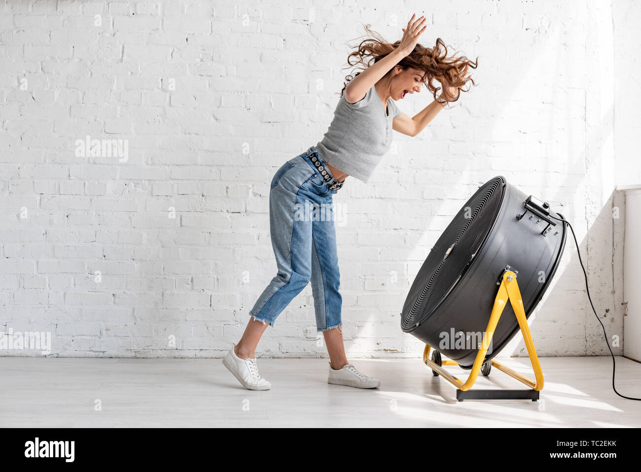 Excitée jeune femme en jean bleu debout avec les mains posées devant le ventilateur de soufflage Banque D'Images