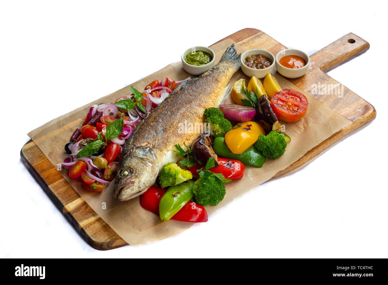 Close up de méditerranée grillé poisson entier basse en bois naturel conseil isolé lumière arrière légumes rôti accompagné de pommes mélanger les tomates cerise basilic Banque D'Images