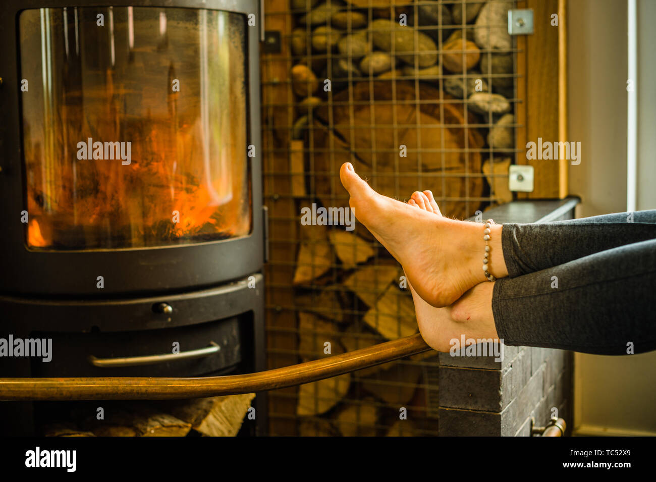 Détail d'une jeune femme portant un bracelet de cheville, réchauffement climatique ses pieds contre un grand feu de bois poêle en fonte. UK Banque D'Images
