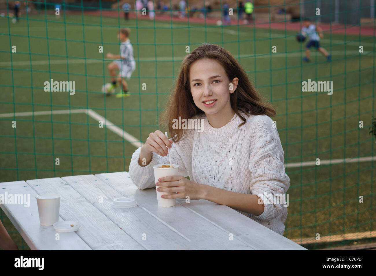 High school student boire du café sur la terrasse d'un café de l'école. Elle sourit et pose pour appareil photo. Terrain de football de l'arrière-plan. Temps pour Banque D'Images