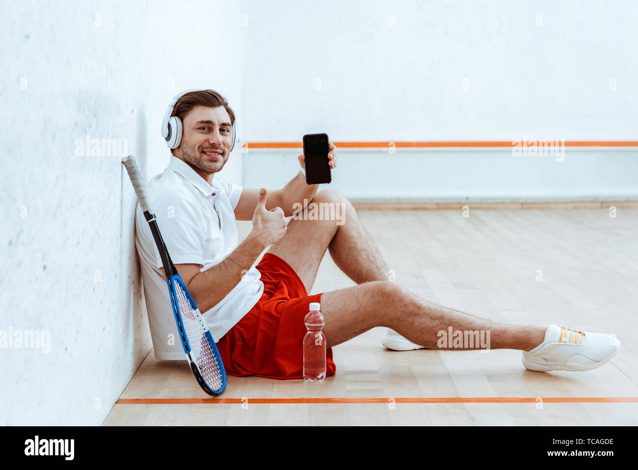 Joueuse de squash dans les écouteurs showing thumb up et holding Smartphone avec écran vide Banque D'Images