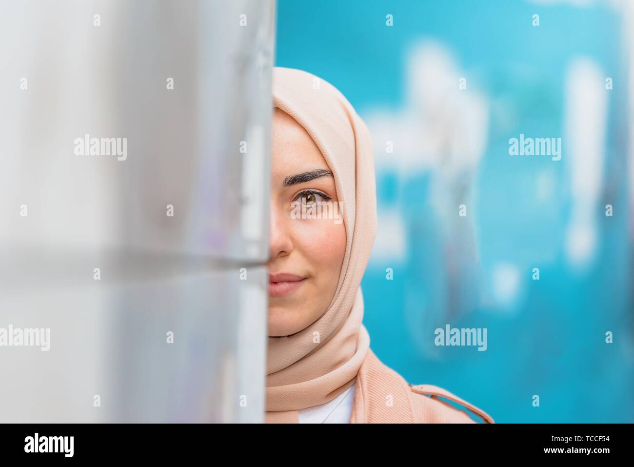 Belle femme musulmane en foulard et des vêtements modernes à la mode se cache la moitié de visage au mur avec fond bleu. Banque D'Images