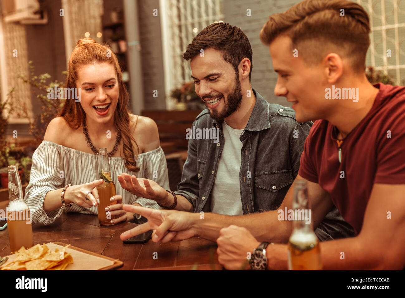 Des boissons avec des amis. Rousse charmante jeune femme séduisante et deux gars attrayant rayonnant heureux assis au bar avec boissons et s'amusant Banque D'Images