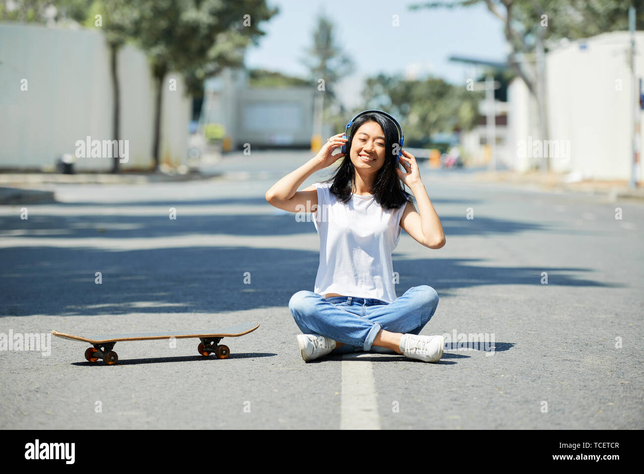 Assez heureux Asian teenage girl sitting on route à côté de skateboard et écouter de la musique dans ses écouteurs Banque D'Images