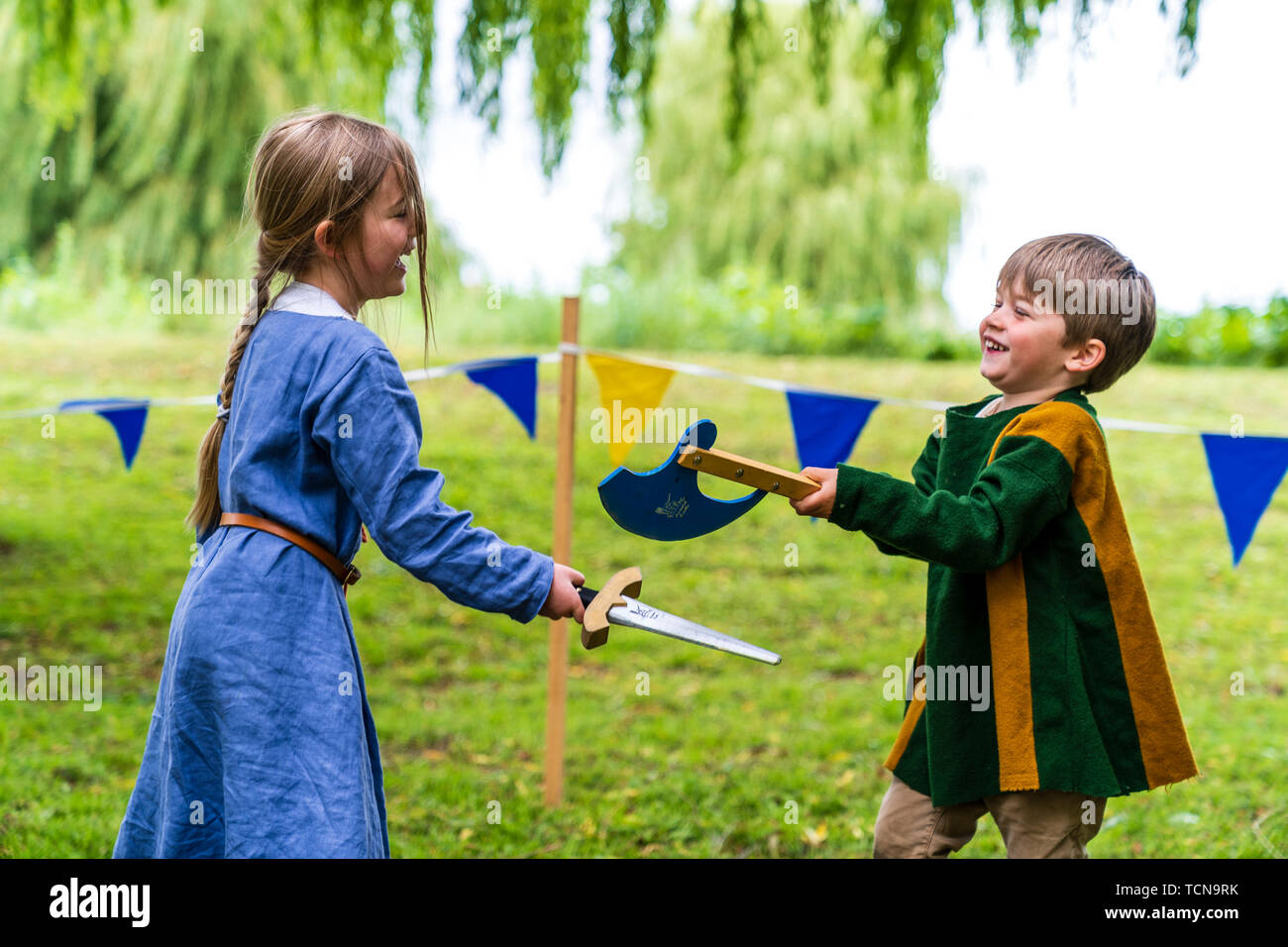 Le week-end médiéval. Portrait de deux jeunes enfants, garçon, 4-5 ans, et une fille, 5-6 ans, vêtu de vêtements médiévaux et des combats à l'épée et espièglerie ax dans l'herbe avec saule derrière. Banque D'Images