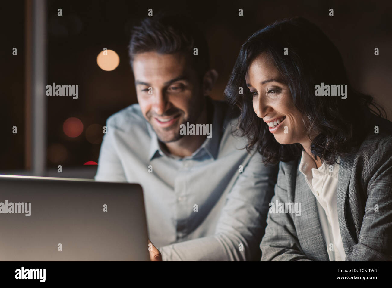 Deux smiling businesspeople assis à un bureau à l'aide d'un ordinateur portable tout en travaillant ensemble dans un bureau, les heures supplémentaires de nuit Banque D'Images