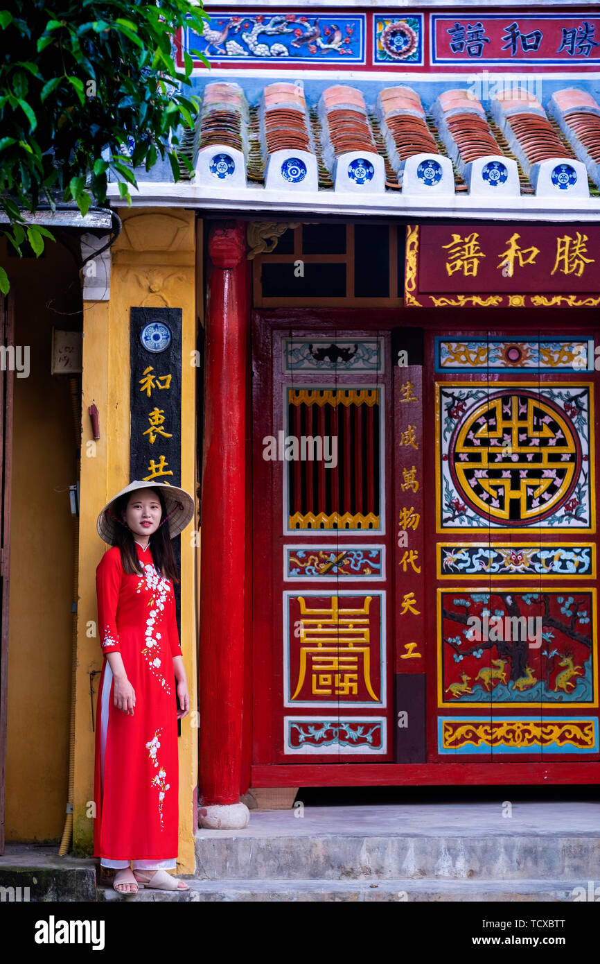 Un jeune Vietnamien femme portant une robe traditionnelle Ao Dai debout à l'extérieur et un temple dans le centre-ville historique, Hoi An, Vietnam, Indochine, Sou Banque D'Images