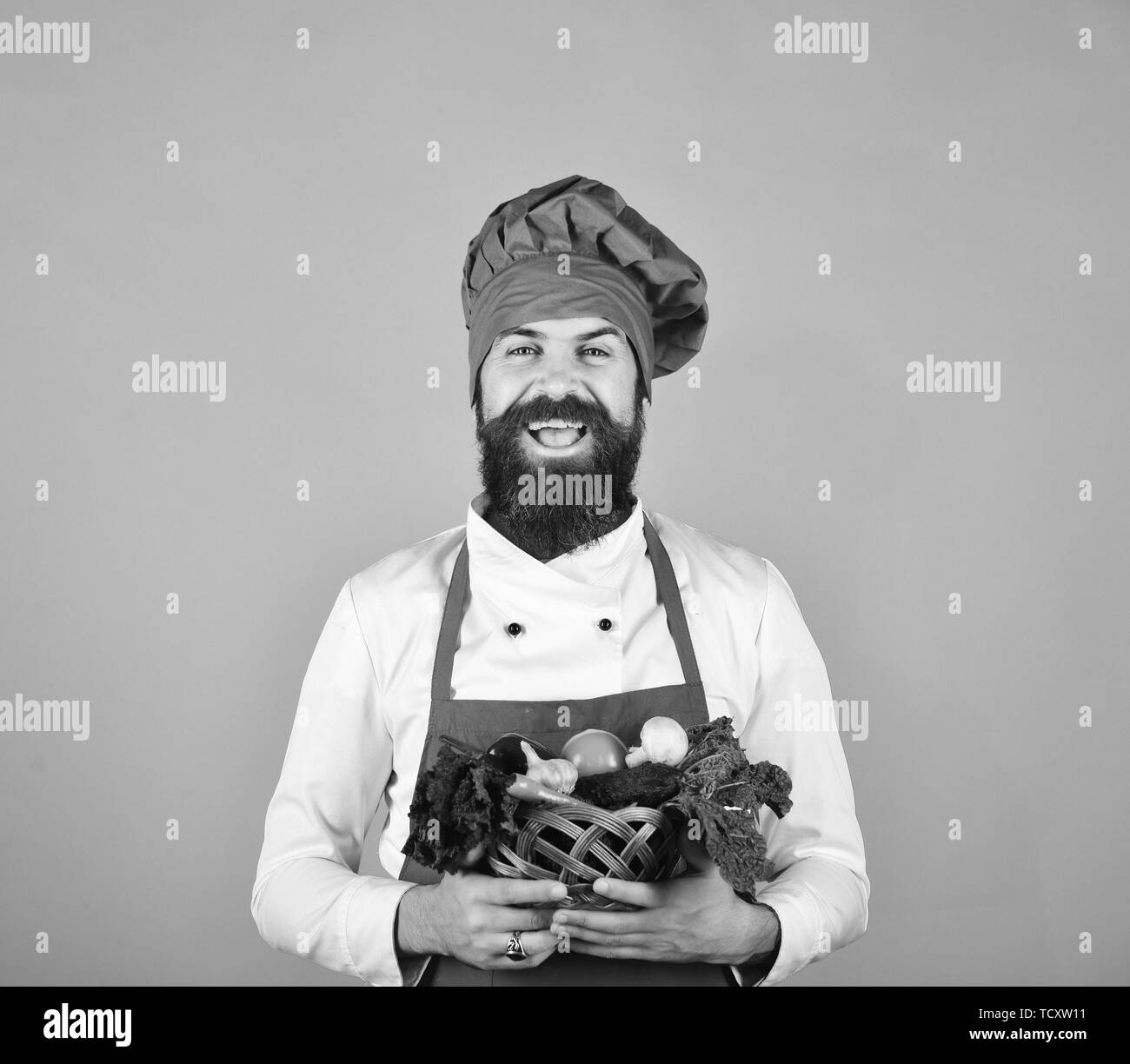 Chef détient la laitue, la tomate, le poivron et les champignons. Concept de cuisine saine. Homme à barbe sur fond rouge. Cook avec visage rieur en bourgogne est titulaire d'uniforme des légumes dans un bol en osier. Banque D'Images