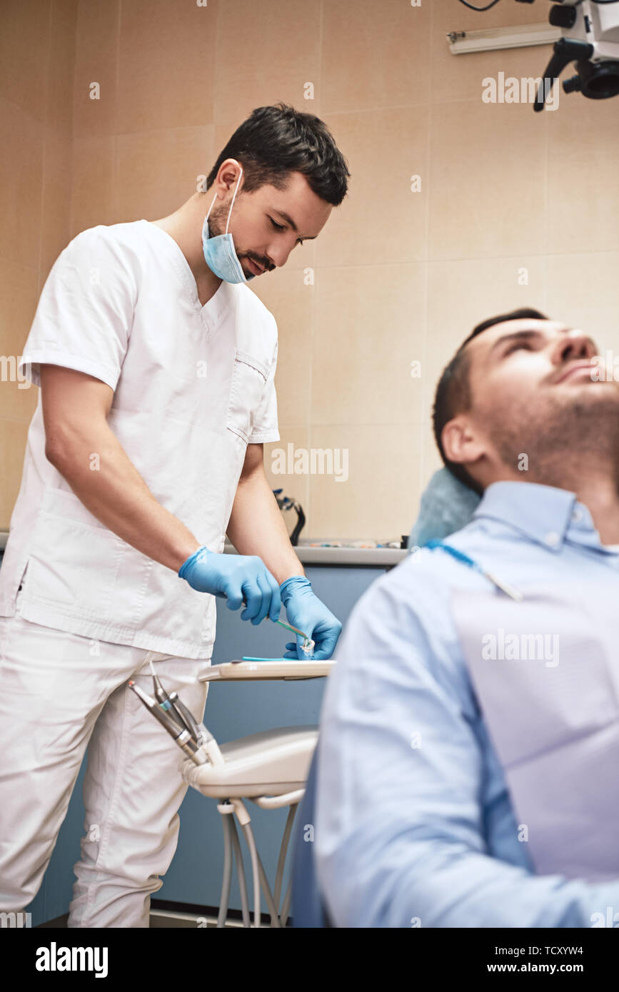 Dark-haired barbu portant des hygiénistes dentaires uniforme blanc est prêt à contrôler les dents du patient. Banque D'Images