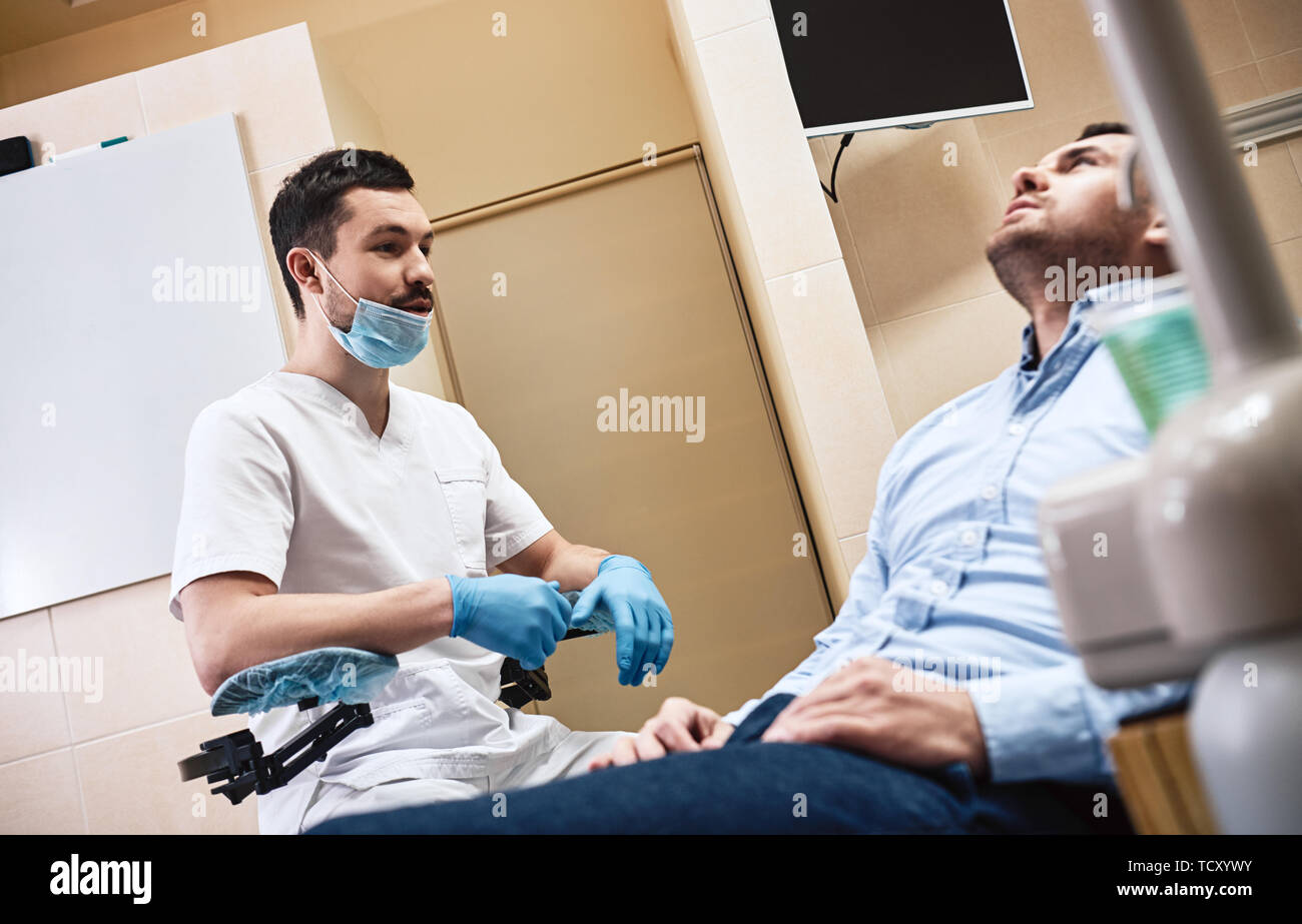 Dark-haired barbu portant des hygiénistes dentaires uniforme blanc est prêt à contrôler les dents du patient. Il est assis près de fauteuil dentaire parle à son patient. Sid Banque D'Images