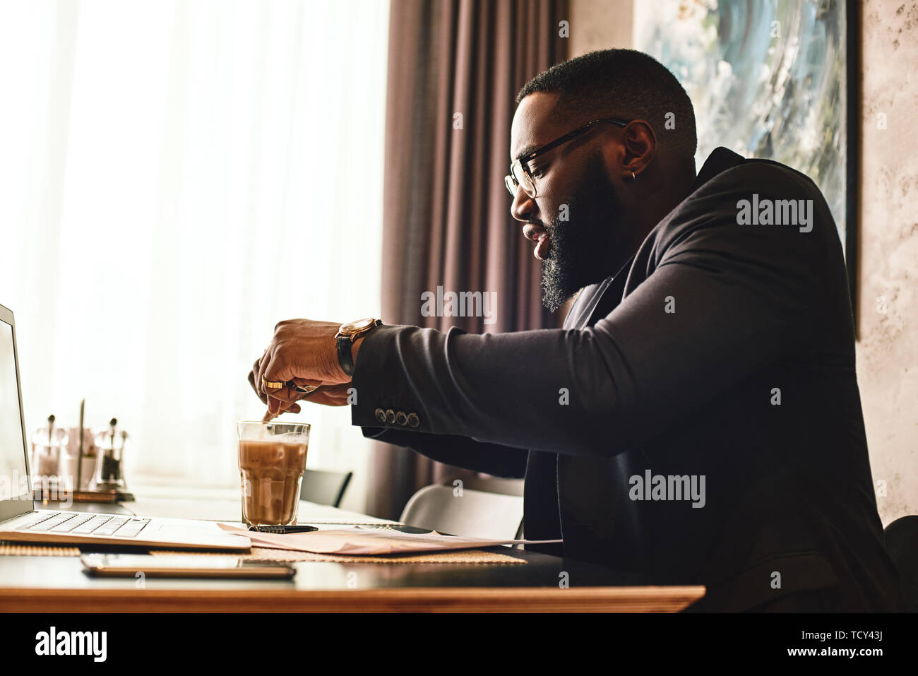 Portrait d'homme à la peau sombre dans le noir-bleu et verres assis à la table, à l'intérieur, de boire du café. Les jeunes entrepreneurs africains met s Banque D'Images