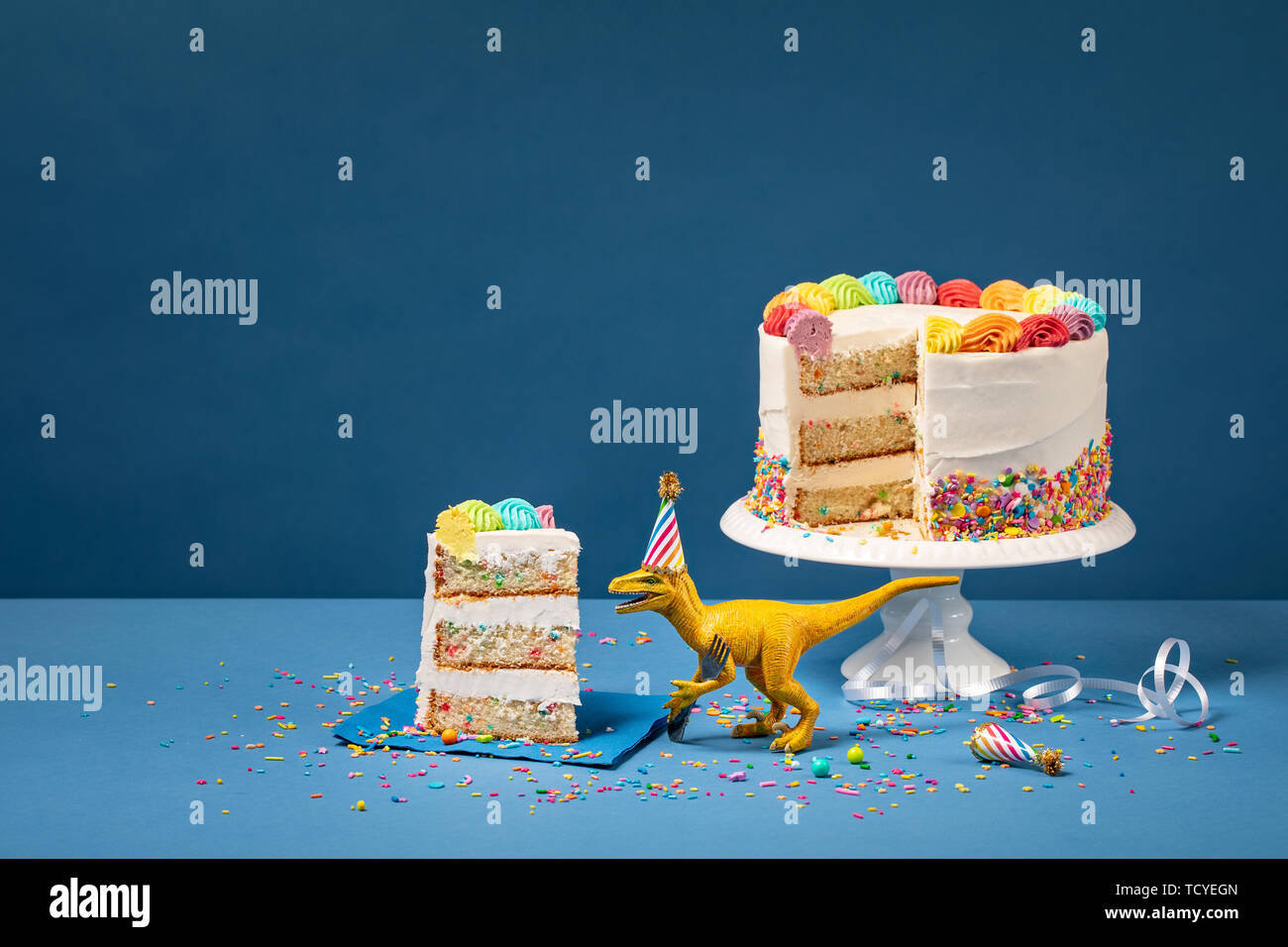 Hungry toy dinosaur portant un chapeau et tenant une fourche à côté d'un gâteau d'anniversaire sur un fond bleu Banque D'Images