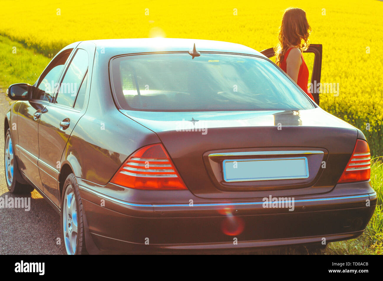 Belle jeune fille dans une robe rouge close up en sortant de la voiture près de la zone jaune avec fleurs de radis et de la lumière du soleil. Banque D'Images