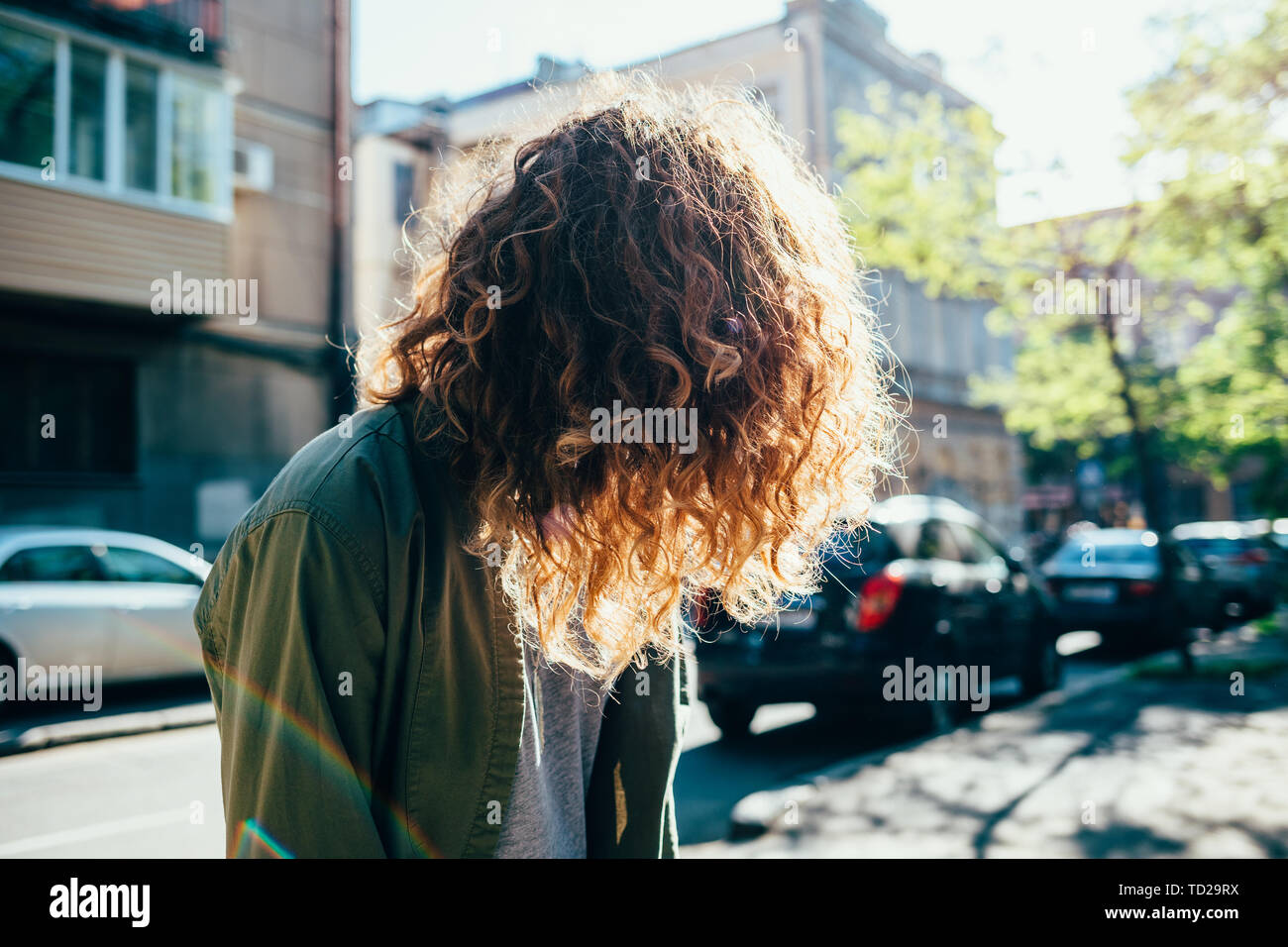 Méconnaissable fille aux cheveux naturellement bouclés debout sur rue près de route avec le trafic au jour d'été ensoleillé. Banque D'Images