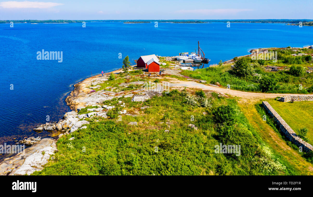 Hasslo, Suède - juin 3, 2019 : pêche rouge cabanes et un petit port dans un très joli paysage côtier. Les gens, chien et chariot visible par les hangars. Banque D'Images