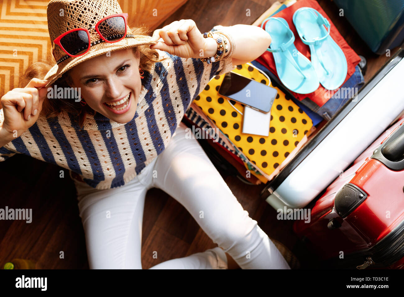 Vue supérieure de smiling femme élégante dans un pantalon blanc et Chemisier rayé avec le souper chapeau et des lunettes de près d'une valise de voyage dans la vie moderne ro Banque D'Images