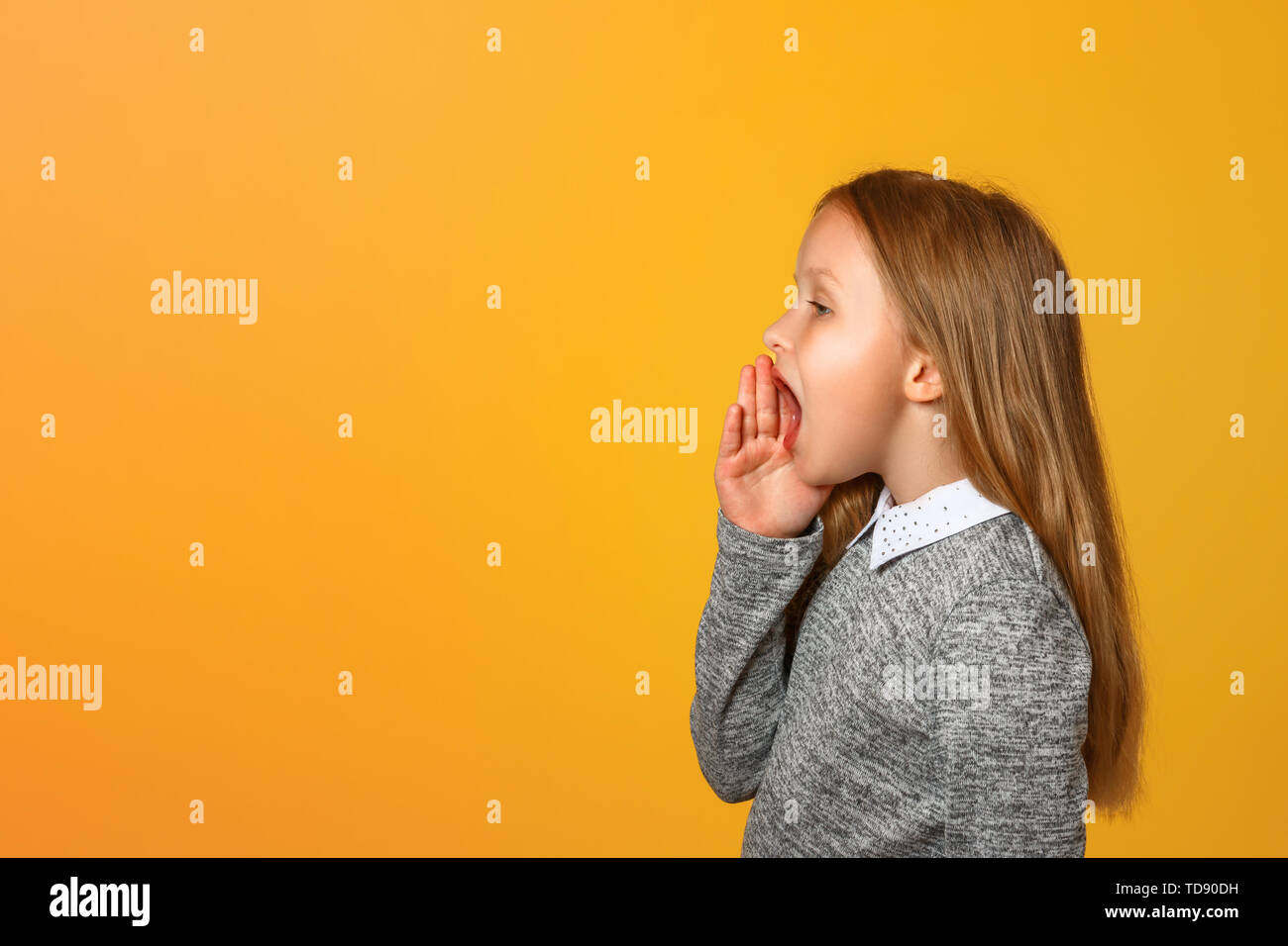 La petite fille crie très fort sur le côté avec une main sur sa bouche. Close-up. Fond jaune. Copier l'espace. Banque D'Images