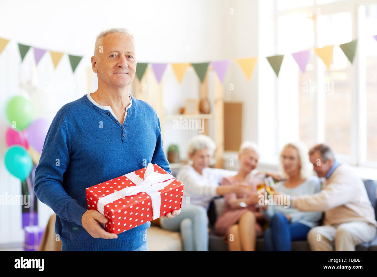 Portrait de l'homme anniversaire beau contenu dans pull bleu debout dans la salle de séjour décorée pour anniversaire et holding gift box pendant que ses amis Banque D'Images