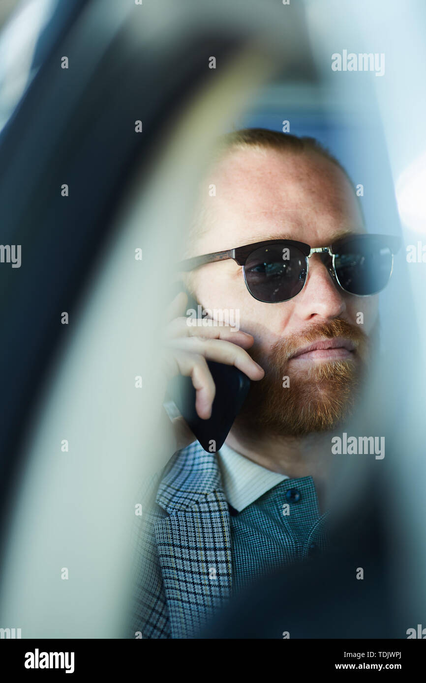 Réfléchis sérieusement entrepreneur barbu à lunettes assis à l'intérieur de la voiture et résoudre les questions de travail sur le téléphone Banque D'Images