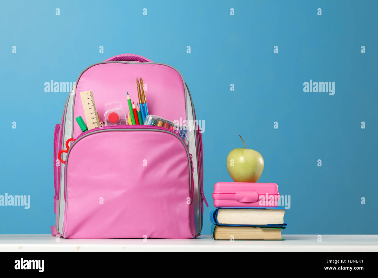 Ensemble des élèves. Sac à dos rose avec modèle, une pile de livres, une  boîte à lunch, une pomme sur la table sur un fond bleu. Retour à l'école  Photo Stock -