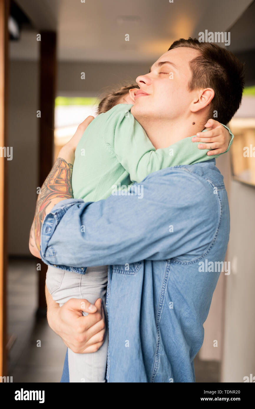 Hipster jeune père aimant calme avec tattoo sur le bras en gardant les yeux fermés tout en serrant sa fille adorable et réconfortant Banque D'Images