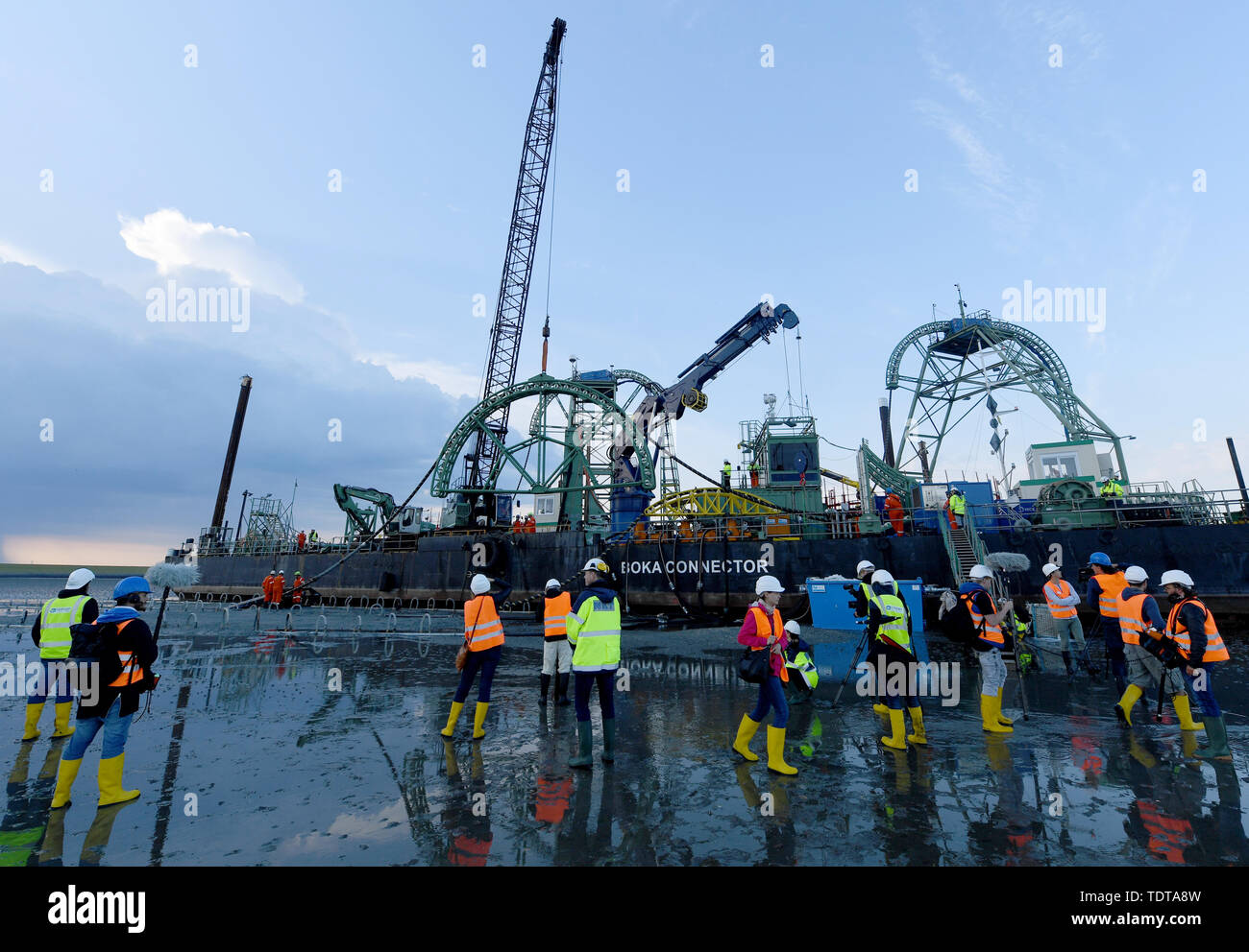 19 juin 2019, le Schleswig-Holstein, Büsum : La fin d'un câble sous-marin 516 kilomètre de long est tiré à terre par le navire câblier "Connecteur Boka' dans la boue sous la digue. Les représentants de la presse se tenir à une distance de sécurité en face de lui avec des gilets haute visibilité. NordLink est le nom de la ligne électrique que pour la première fois va se connecter directement les marchés de l'électricité de l'Allemagne et la Norvège et servira d'échange entre l'allemand et de l'énergie éolienne l'énergie hydraulique norvégien. D'ici la fin de 2019, le travail final aurait dû être terminée. Photo : Carsten Rehder/dpa Banque D'Images