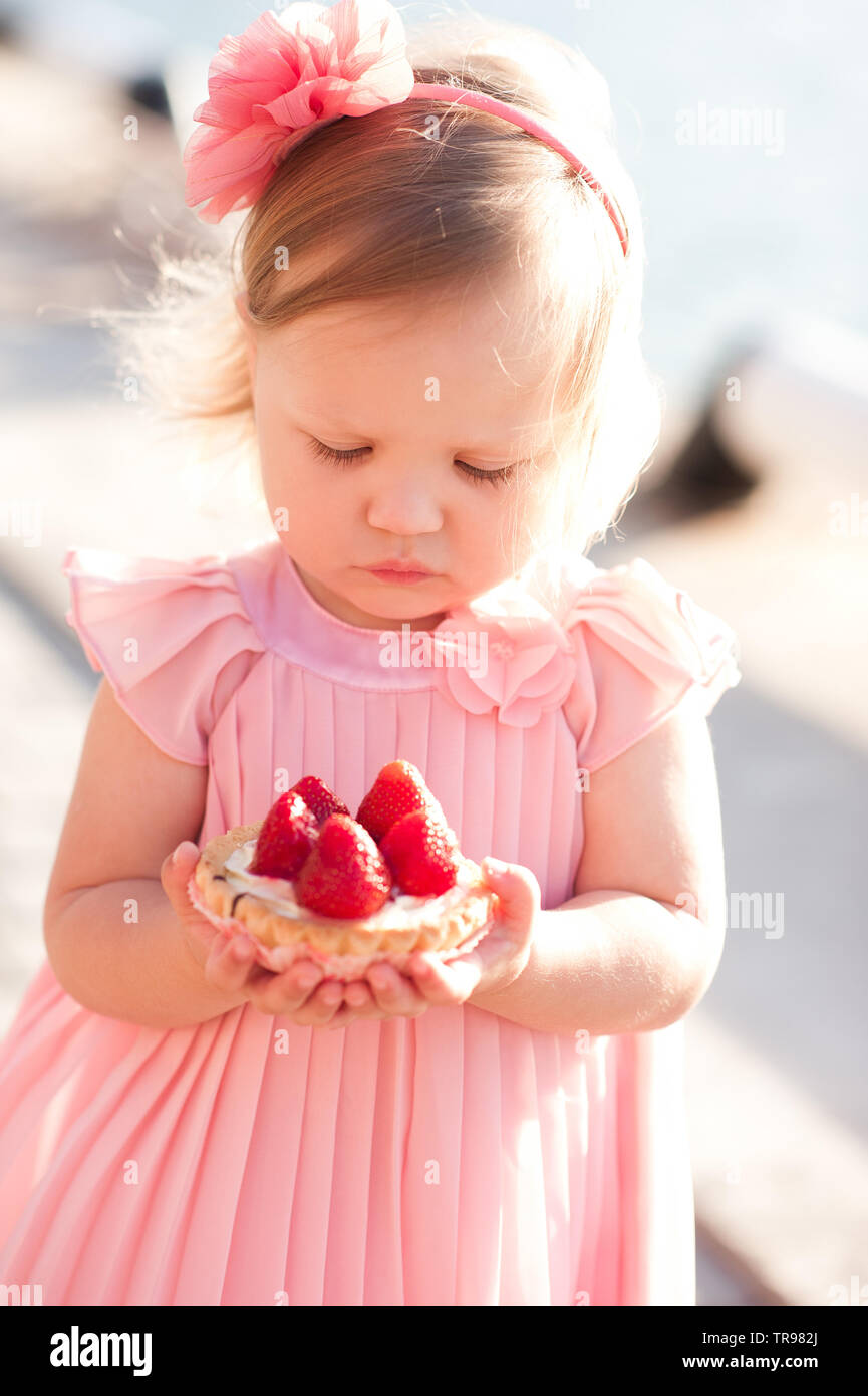 Mignon bébé fille 2-3 ans holding strawberry cake à l'extérieur. Porter  robe rose élégant. Regardant vers le bas. L'enfance Photo Stock - Alamy