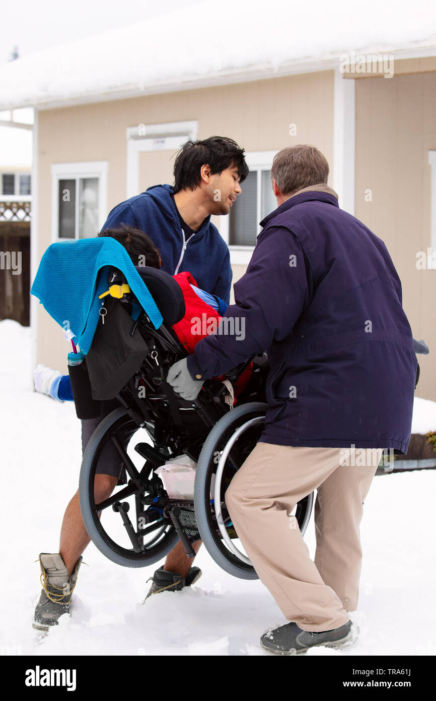 Deux hommes de soulever ou de transporter un fauteuil roulant avec les jeunes personnes handicapées garçon assis, sur la neige en hiver Banque D'Images