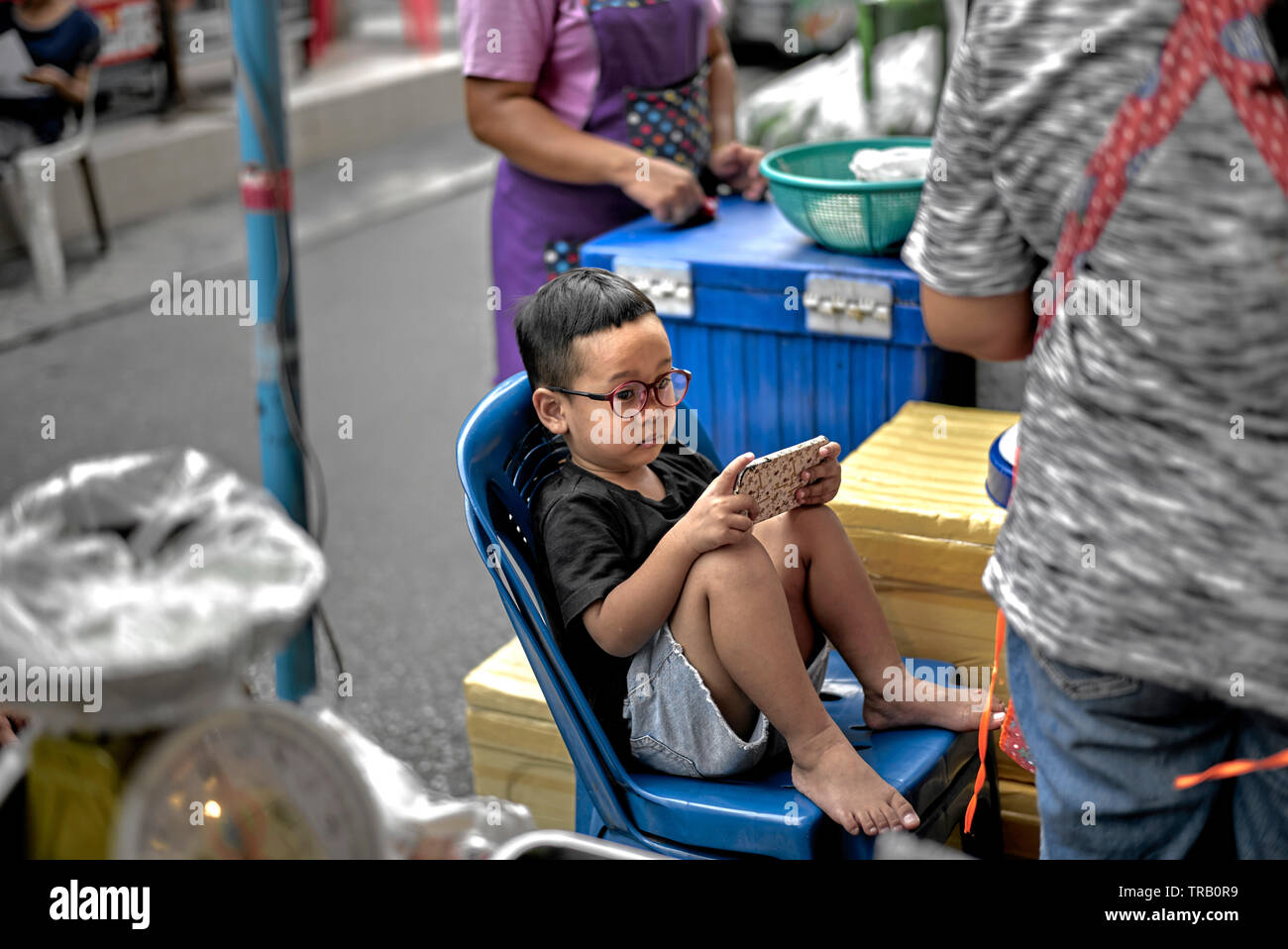 Lunettes pour enfants. Enfant portant des lunettes jouant sur un smartphone dans une rue thaïlandaise Banque D'Images