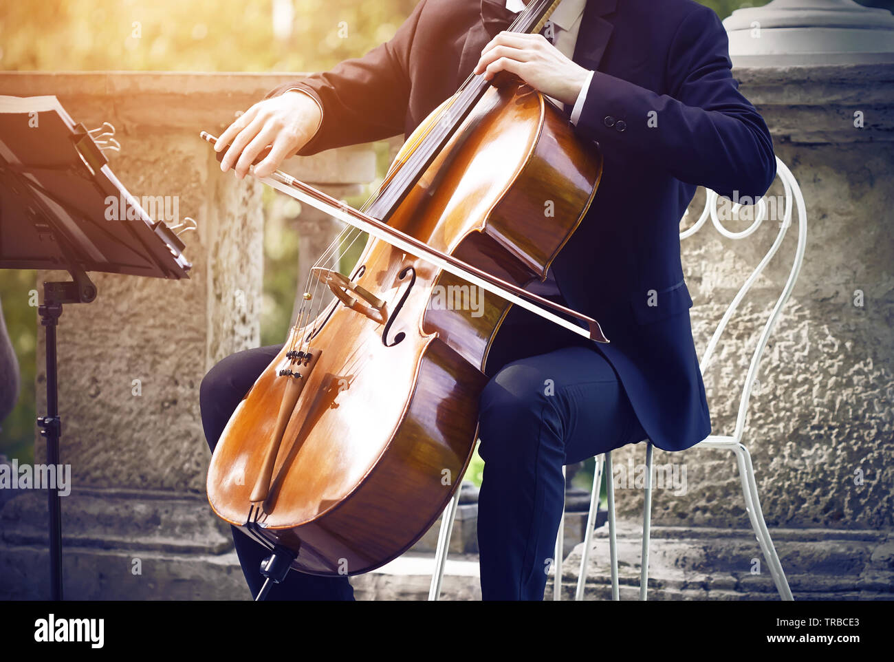 Musicien dans un costume noir assis sur un fauteuil élégant blanc sur la terrasse dans le parc et jouer de la musique classique sur l'instrument bow-string - violoncelle. Banque D'Images