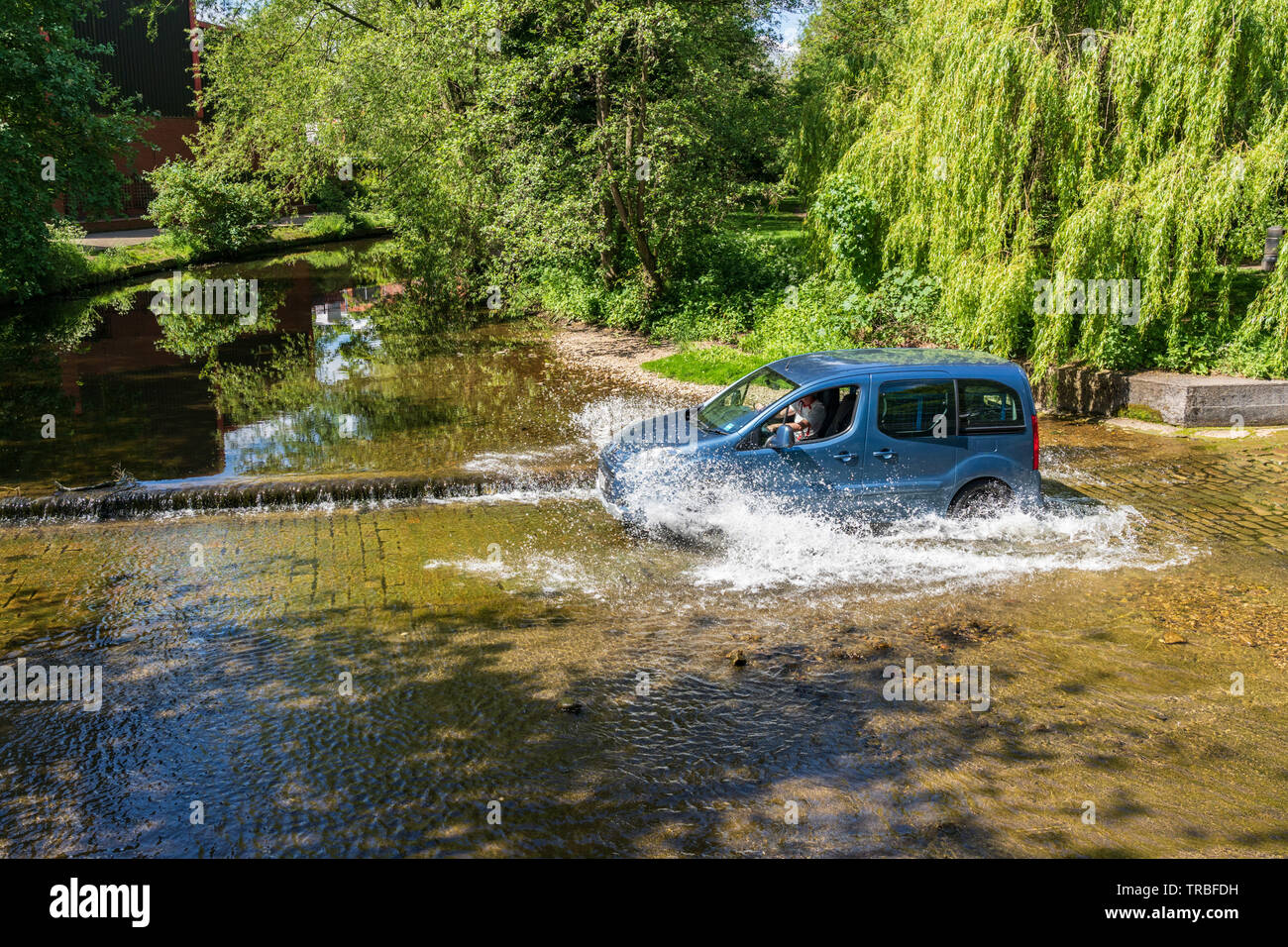 Les éclaboussures d'eau sont visibles le long de la rivière tandis que le petit véhicule bleu traverse une Ford peu profonde, Ripon, North Yorkshire, Angleterre, Royaume-Uni. Banque D'Images
