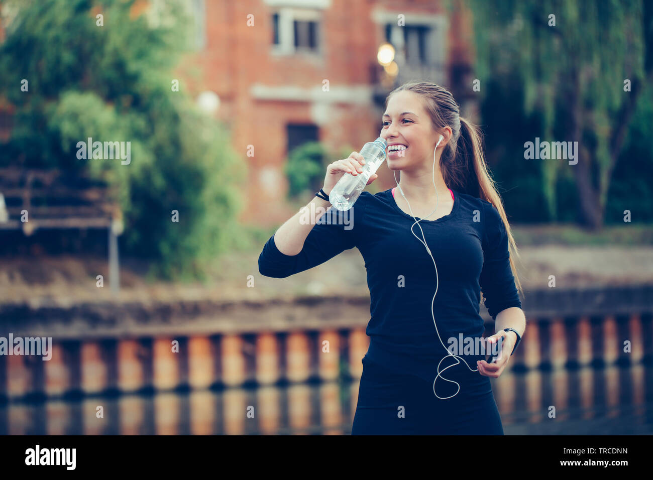 Coureuse debout près de la rivière tenant une bouteille d'eau. Femme de remise en forme en faisant une pause après l'exécution de l'entraînement. Banque D'Images